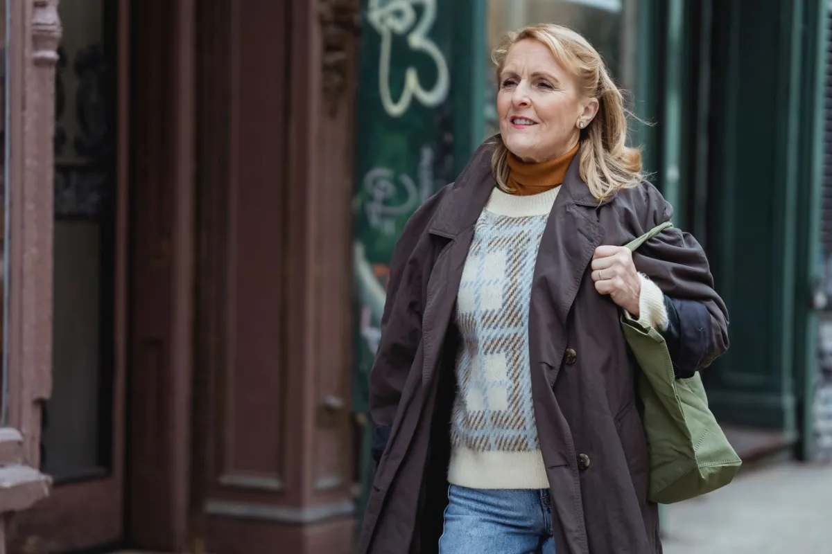 Mature female in checkered sweater and coat with bag looking away while strolling on walkway in city